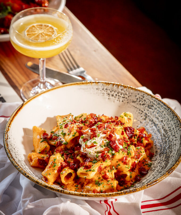 Bowl of rigatoni pasta in a spicy cream sauce topped with crispy bacon, burrata, and fresh chives, served alongside a yellow citrus cocktail in a coupe glass on a wooden table