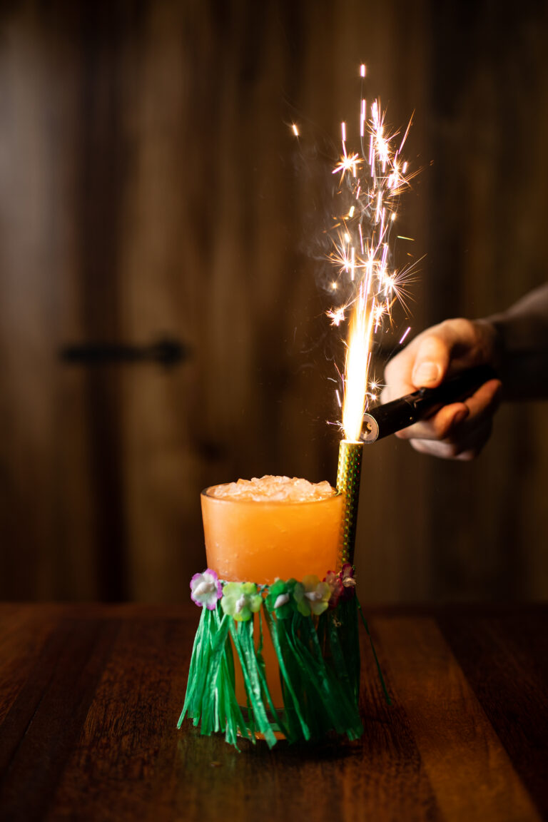 Tropical bourbon punch cocktail with a sparkler and hula skirt garnish being lit.