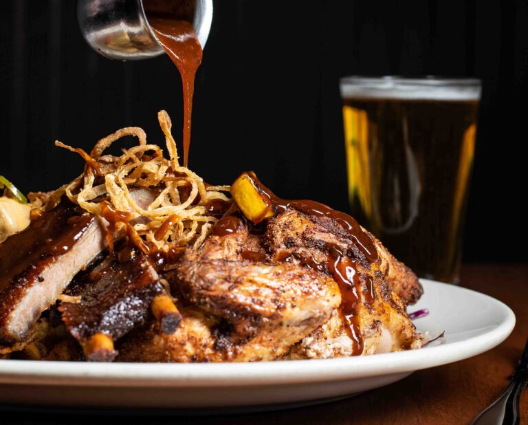 Chef pouring rich house sauce over a platter of smoked meats topped with crispy onions at Sharps RoastHouse.