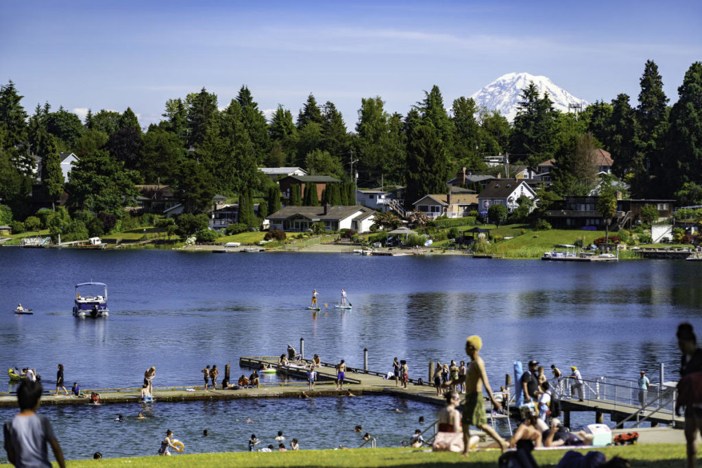 Photo of beautiful Angle Lake. It is a bright summer day and there are many people enjoying lakeside activities like sunbathing, swimming, stand-up paddle boarding, and boating. Mount Rainier can be seen in the distance.