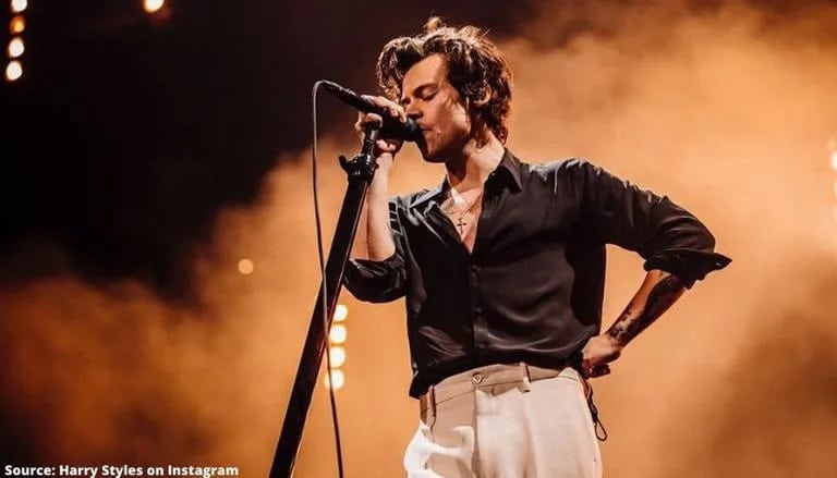 A photo of Harry Styles performing on stage. He has his eyes closed as he sings into a microphone in front of an orange cloud of smoke.