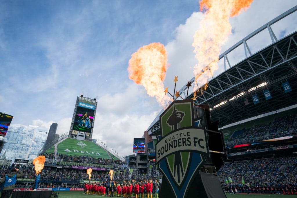 Photo of Century Link Field. The stadium is full of fans as two soccer teams prepare to play. There is a display in the foreground that says "Seattle Sounders FC" with flames erupting from the top.