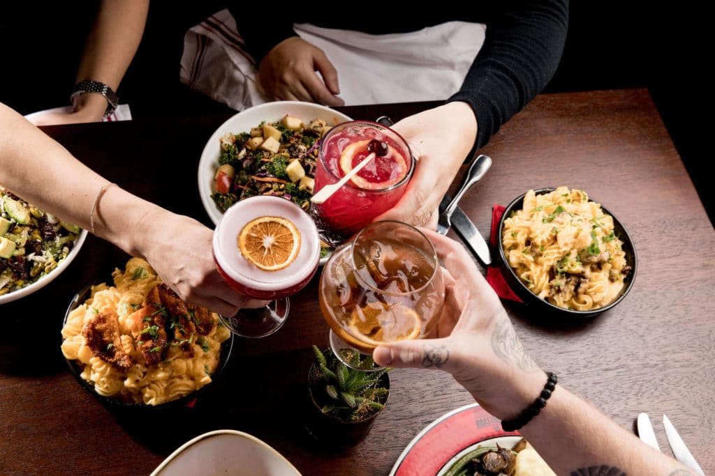 A group of three friends cheers over a delicious looking meal at Sharps Roasthouse. They are each holding a unique and yummy cocktail.