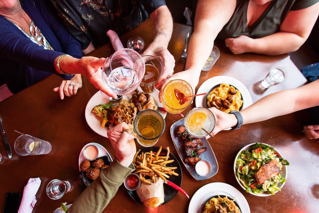 A group of 5 people are reaching in for a "cheers" over a table of delicious looking food.