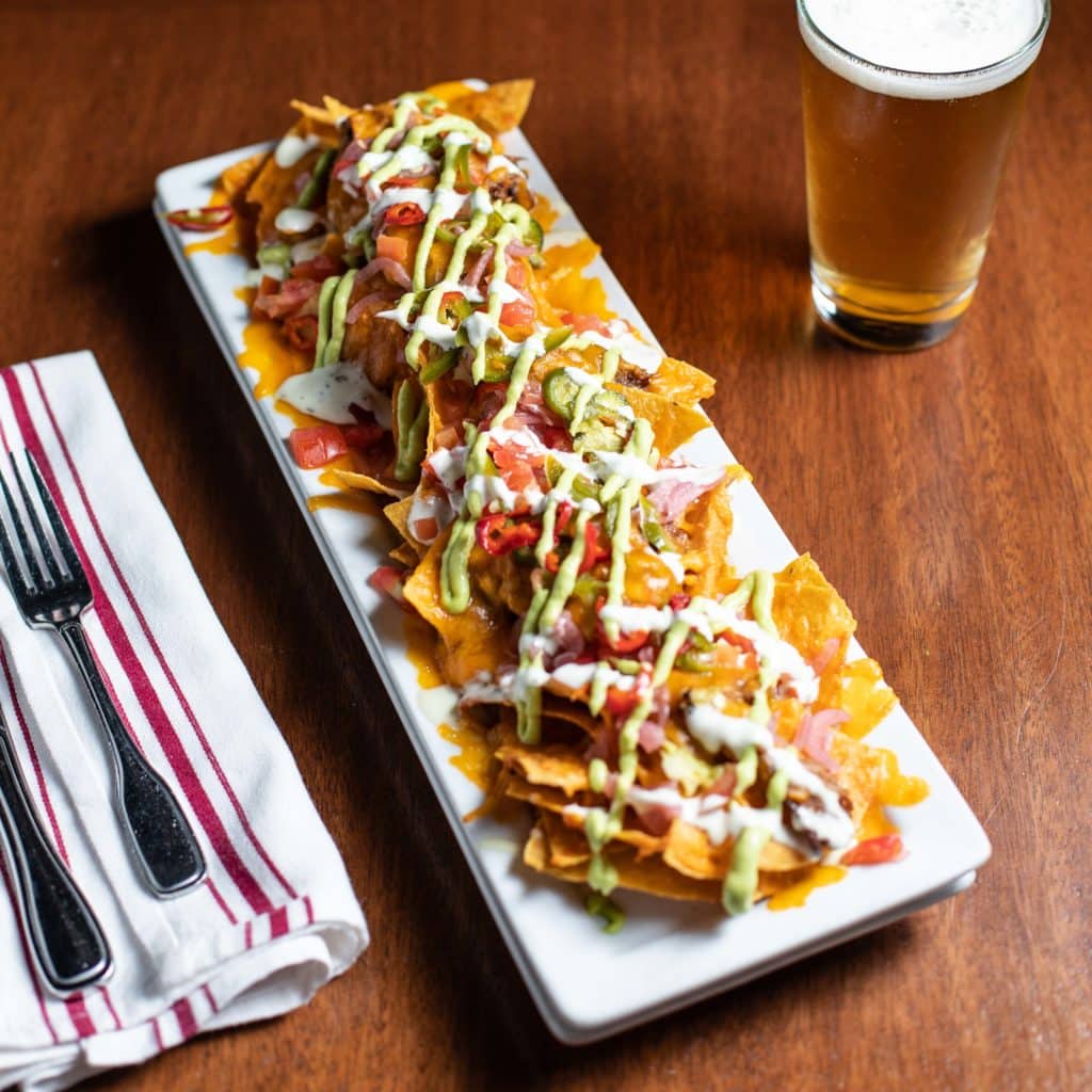 Picture of Bar Boss Bourbon™ BBQ Pork Nachos on a wooden table next to a cold beer. 
The nachos have scratch corn tortilla chips, cheddar cheese, tomatoes, pickled onions, Fresno chiles, avocado salsa, crema, and Bar Boss Bourbon BBQ sauce