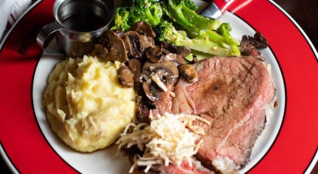 Picture of a delicious looking meal. There is a juicy prime rib with fresh horseradish on top, a side of mashed potatoes, sautéed mushrooms, and charred broccoli.