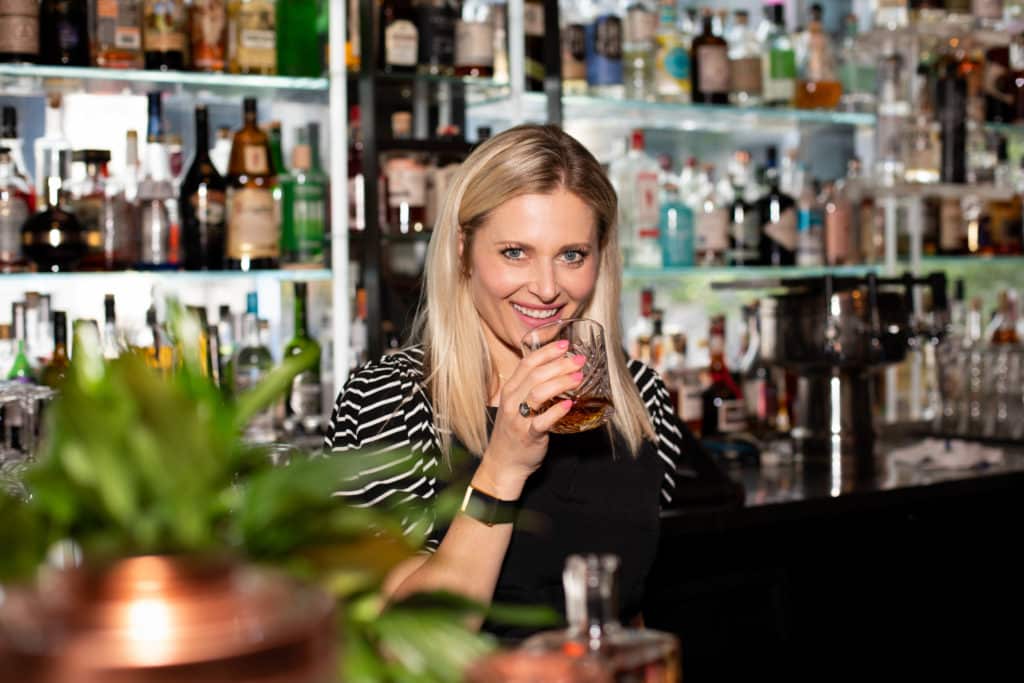 Portrait of Sharps Roasthouse Owner and Operator, Missy Firnstahl-Claridge. 
A blonde woman is standing in front of a well-stocked bar holding an etched glass partially filled with Bourbon in her right hand. She is smiling and looking pleasant.
