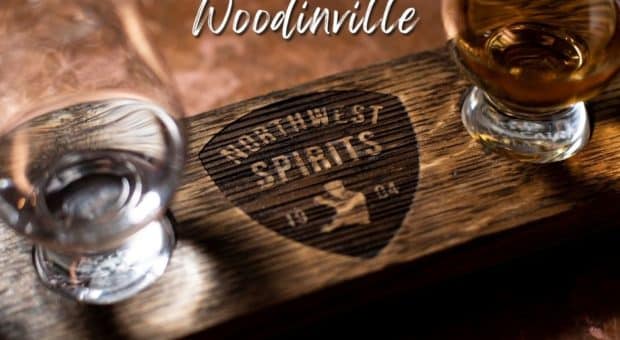 A wooden spirit-tastings board with "Northwest Spirits" carved into it sits atop a copper bar top. There are 2 glasses on either side of the engraving - a whiskey and a vodka. There is a text overlay on the top of the image that reads "See you soon, Woodinville".