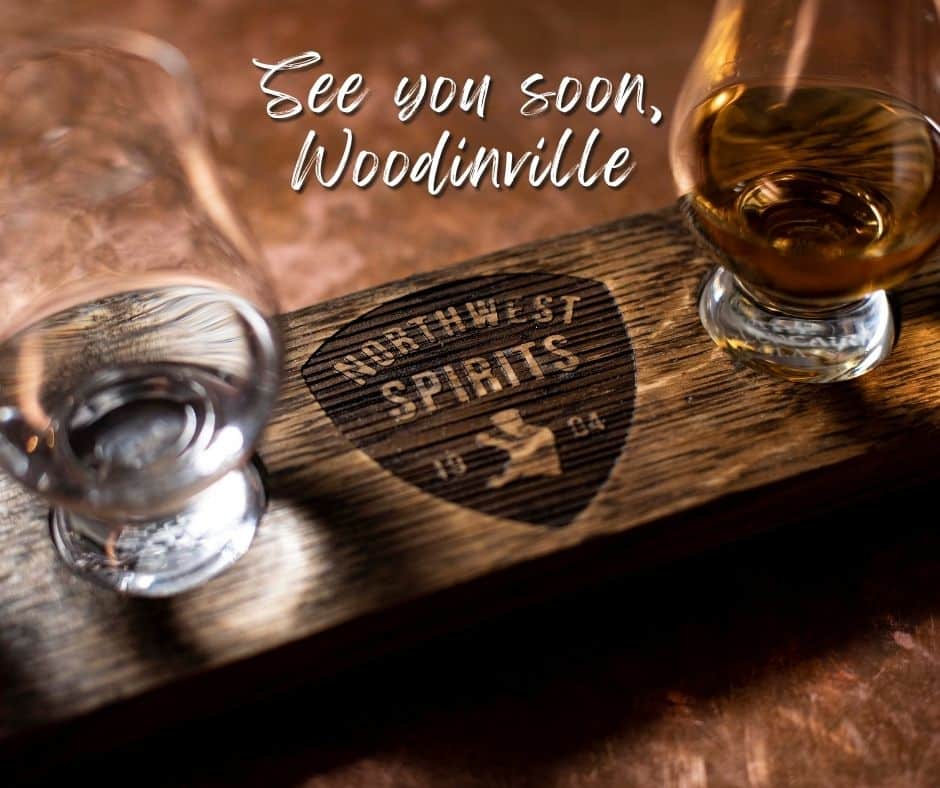 A wooden spirit-tastings board with "Northwest Spirits" carved into it sits atop a copper bar top. There are 2 glasses on either side of the engraving - a whiskey and a vodka. There is a text overlay on the top of the image that reads "See you soon, Woodinville". 