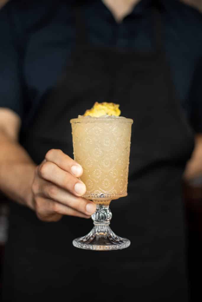 A man’s hand is holding a peach-colored drink in an etched tall glass. The man is wearing a black apron. The drink has a slice of dried pineapple on top.