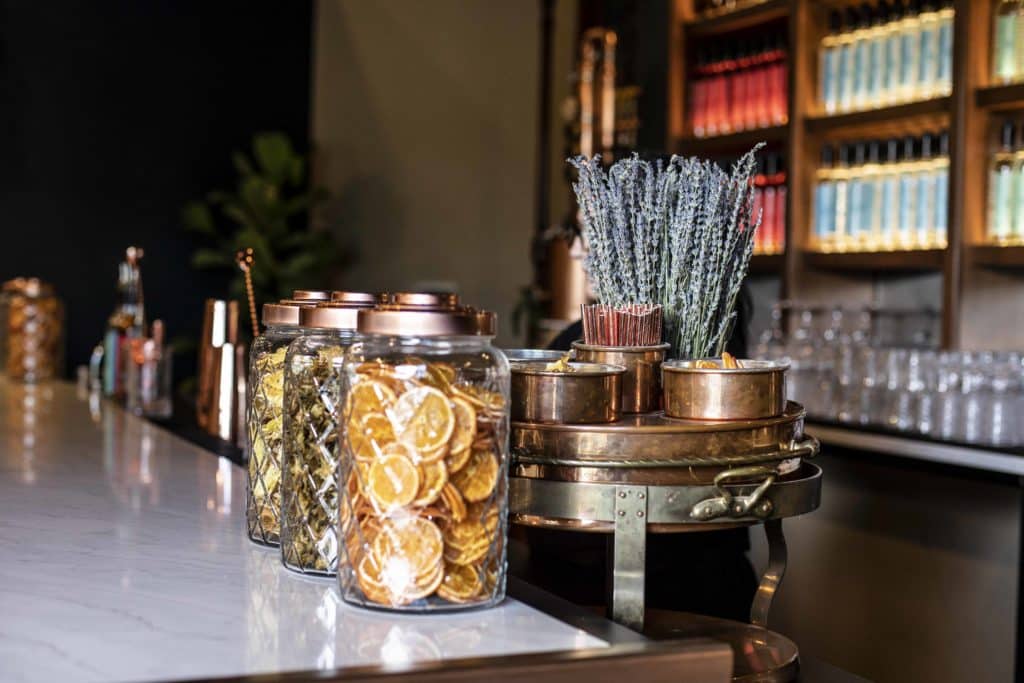 A white bar top sits in front of an impressive selection of liquor bottles lined up neatly on a Mahogany wooden display. The bar as 3 glass jars on top filled with dried fruits. There is a copper container holding various garnishes behind the jars. 