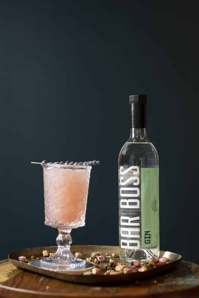 Image of a bottle of Bar Boss Gin sitting on top of a copper tray in front of a dark blue wall. There is a blush pink drink in an etched tall glass next to the bottle. There are dried rose and coriander sitting on the copper tray.