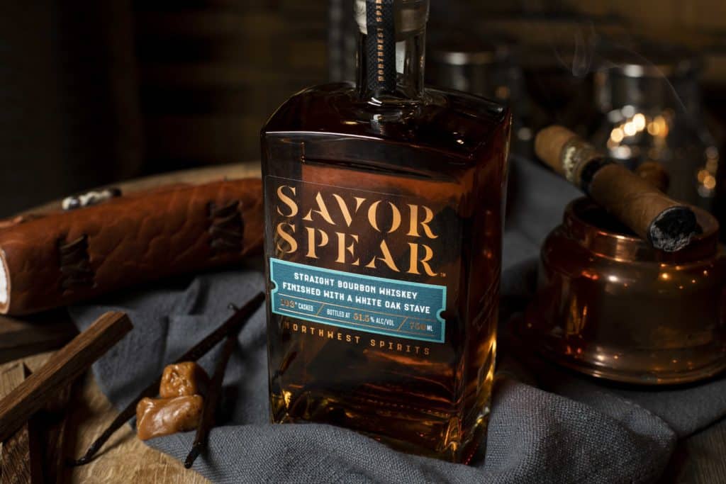 Warm image of a whiskey bottle sitting on a wooden barrel. The Bottle reads “Savor Spear Straight Bourbon Whiskey”. The bottle is sitting atop a blue napkin. The surface around the bottle is adorned with a lit cigar, two soft caramel candies, two vanilla beans, and a leather notebook.  