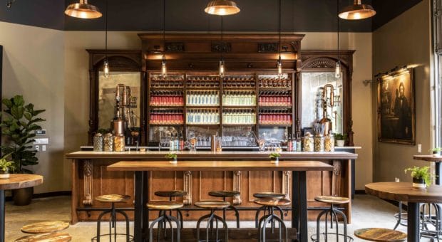 Image of an inviting tasting room. There is an impressive selection of liquor bottles lined up neatly on a Mahogany wooden display behind a white bar top. There are a few wooden tables with wooden stools. There is an industrial, yet warm feeling in the space.