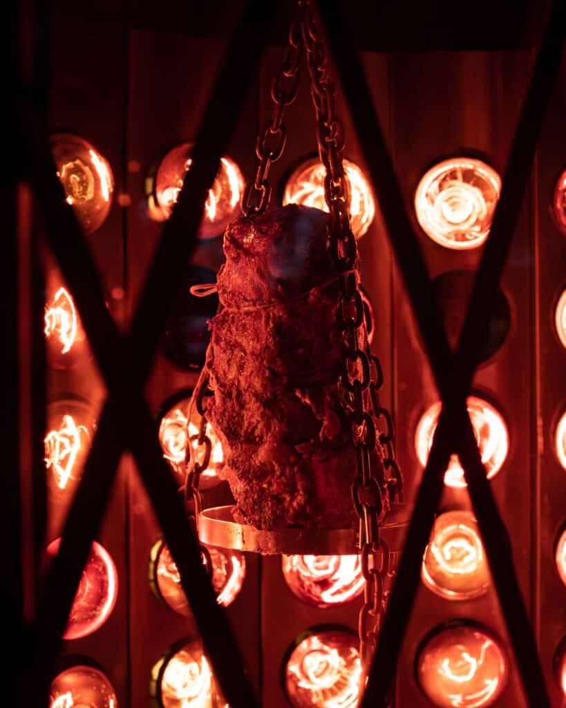 Image of the Radiant Roaster at Sharps RoastHouse. A large cut of Prime Rib is suspended in the center of the roasting case, tied with twine to a metal rack with chains. The image is glowing red from the roaster light bulbs, which line the back of the case.