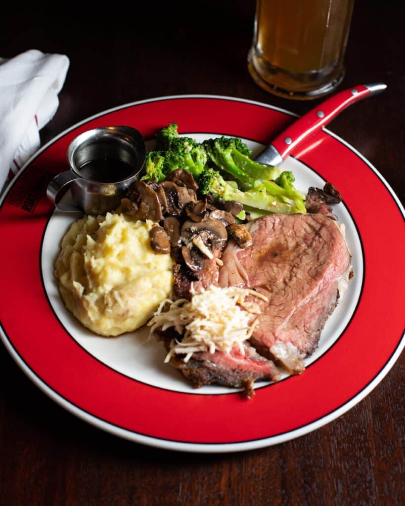 Image of a delicious looking meal consisting of a medium rare slice of Prime Rib topped with fresh horseradish and sautéed mushrooms. On the side there is creamy mashed potatoes and steamed broccoli with a small container of au jus, meant for pouring over the meat.