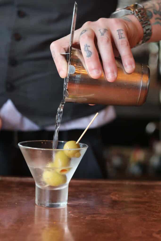 Image of a server with tattoos pouring a vodka martini with 3 olives.