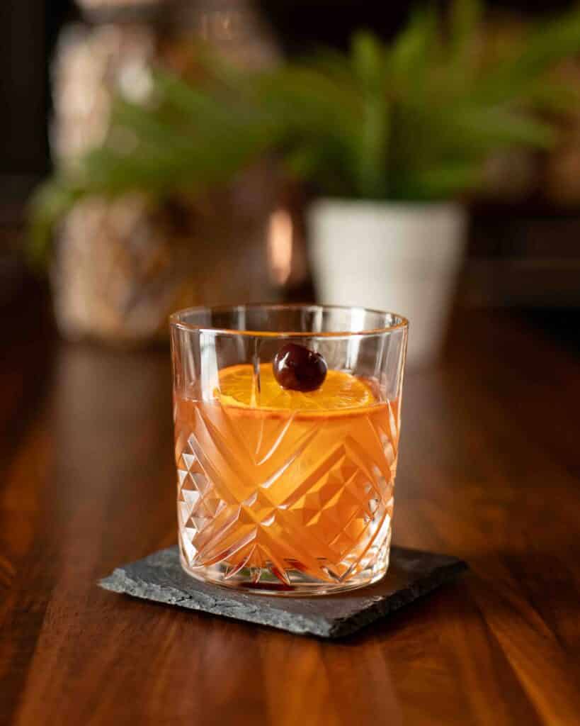 Crystal glass of a Barrel Aged Old Fashioned cocktail garnished with an orange slice and cherry, served on a slate coaster.