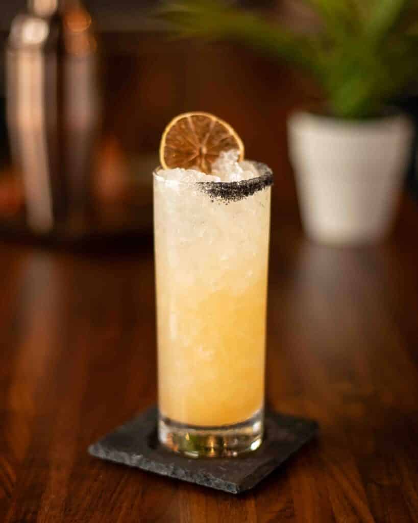 Tall glass of Hot Honey Paloma cocktail with crushed ice, lime wheel garnish, and a black salt rim, served on a dark slate coaster.
