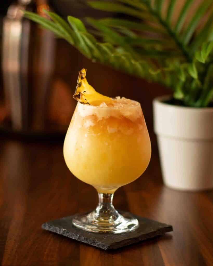 Tropical Painkiller cocktail served in a tulip glass with crushed ice, pineapple wedge garnish, and a dark rum float.