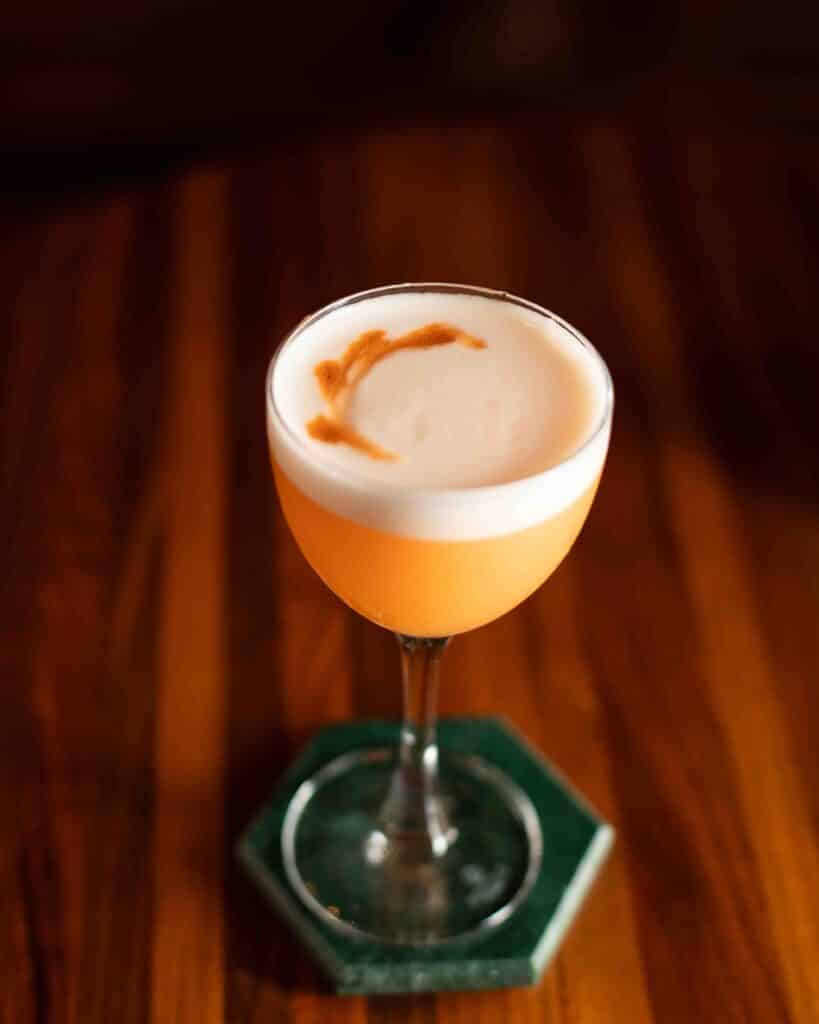 Passion Play cocktail with a foamy egg white top and bitters garnish, served in a coupe glass on a green hexagon coaster.