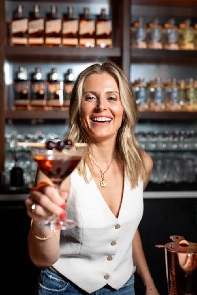 Smiling female owner, Missy Claridge, at Sharps RoastHouse holding up a handcrafted bourbon cocktail garnished with Luxardo cherries, standing in front of a backlit bar shelf featuring Northwest Spirits bourbon and vodka bottles.