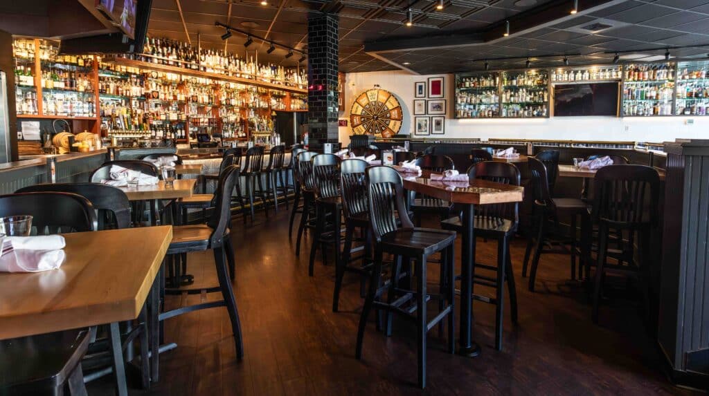 “Interior of a modern, warmly lit restaurant and bar featuring dark wood furnishings, high-top tables, and an extensive wall of spirits behind the bar. A large wooden Wagering Wheel is visible in the background, adding a touch of vintage charm.