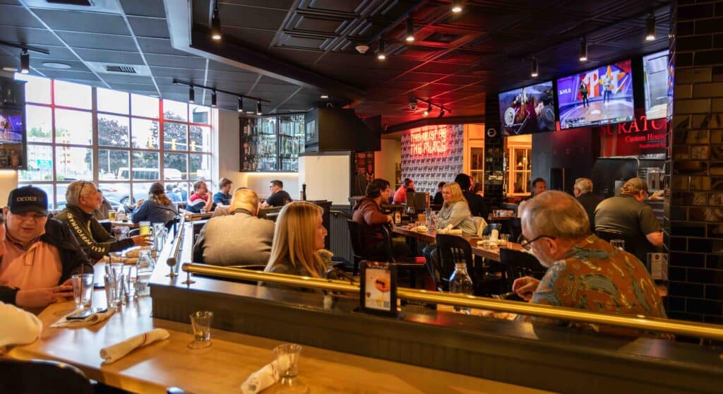nterior of a lively, modern restaurant filled with guests dining and socializing. Large industrial-style windows bring in natural light while multiple flat-screen TVs show sports above the bar. A glowing red neon sign reads “THIS MUST BE THE PLACE” in the background. The atmosphere is warm and welcoming with a mix of high-top and regular tables.